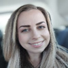 Smiling young woman with long blonde hair sitting indoors, possibly on public transport, conveying a friendly and cheerful expression.