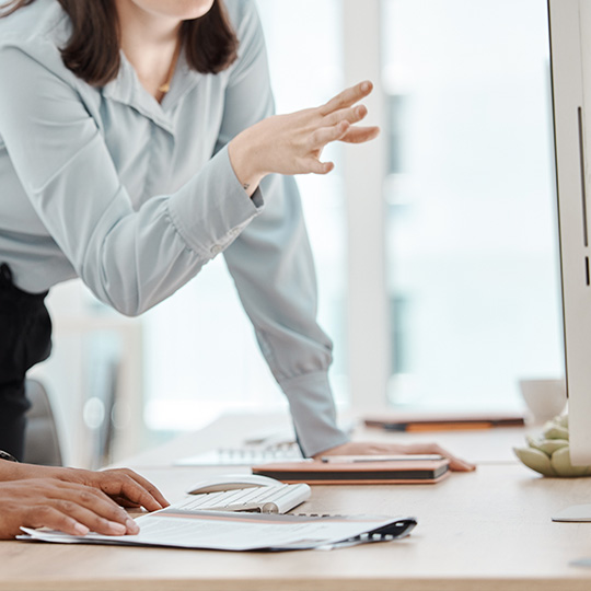 A woman in a light blue blouse is gesturing while discussing something with a person whose hands are visible on a desk with notebooks and a keyboard, in an office setting.