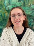 Young woman with long hair wearing glasses and a cozy white sweater, smiling in front of a green lily pad background.