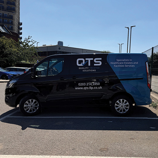 Black commercial van parked in a lot, featuring the logo and contact information for QTS, specialists in healthcare estates and facilities services.