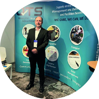 Man in a suit standing in front of a promotional display for QTS, which highlights their services in quality management, facilities services, and asset management.