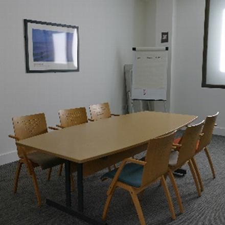 A modern conference room featuring a large wooden table surrounded by six wooden chairs, a whiteboard in the background, and a framed picture on the wall.