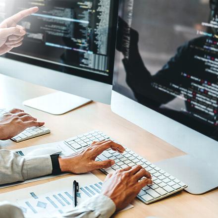 two hands typing on a keyboard in front of computer monitors displaying code, person pointing at the screen, focusing on programming and collaboration