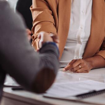 A close-up image of two people engaged in a handshake over a table, with one person wearing a brown blazer and the other in a gray sweater, suggesting a professional meeting or agreement.